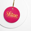 Glossy ceramic Christmas ornament with gold ‘Shine’ lettering on a mystic magenta background, sitting on a white table. Created by Women’s Fiction & Romance Author Juliet Gauvin.