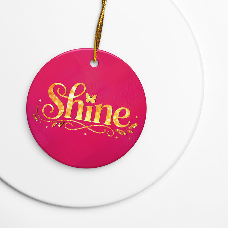 Glossy ceramic Christmas ornament with gold ‘Shine’ lettering on a mystic magenta background, sitting on a white table. Created by Women’s Fiction & Romance Author Juliet Gauvin.