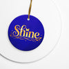 Glossy ceramic Christmas ornament with gold ‘Shine’ lettering on a midnight sapphire background, sitting on a white table. Created by Women’s Fiction & Romance Author Juliet Gauvin.