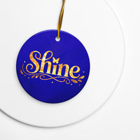 Glossy ceramic Christmas ornament with gold ‘Shine’ lettering on a midnight sapphire background, sitting on a white table. Created by Women’s Fiction & Romance Author Juliet Gauvin.