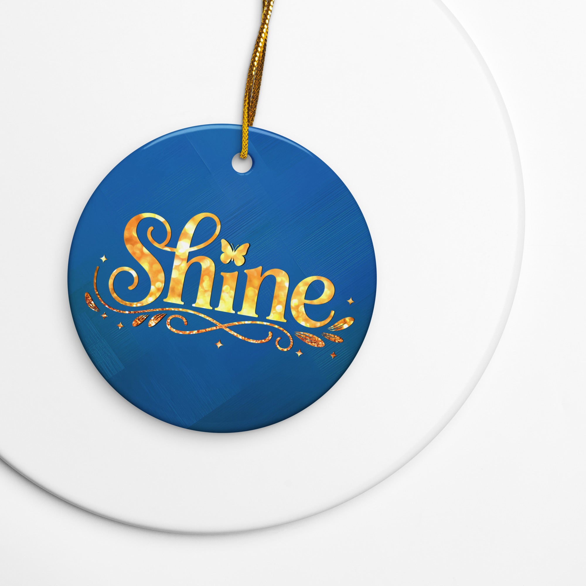 Glossy ceramic Christmas ornament with gold ‘Shine’ lettering on a midnight teal background, sitting on a white table. Created by Women’s Fiction & Romance Author Juliet Gauvin. 