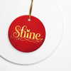 Glossy ceramic Christmas ornament with gold ‘Shine’ lettering on a mags red background, sitting on a white table. Created by Women’s Fiction & Romance Author Juliet Gauvin.